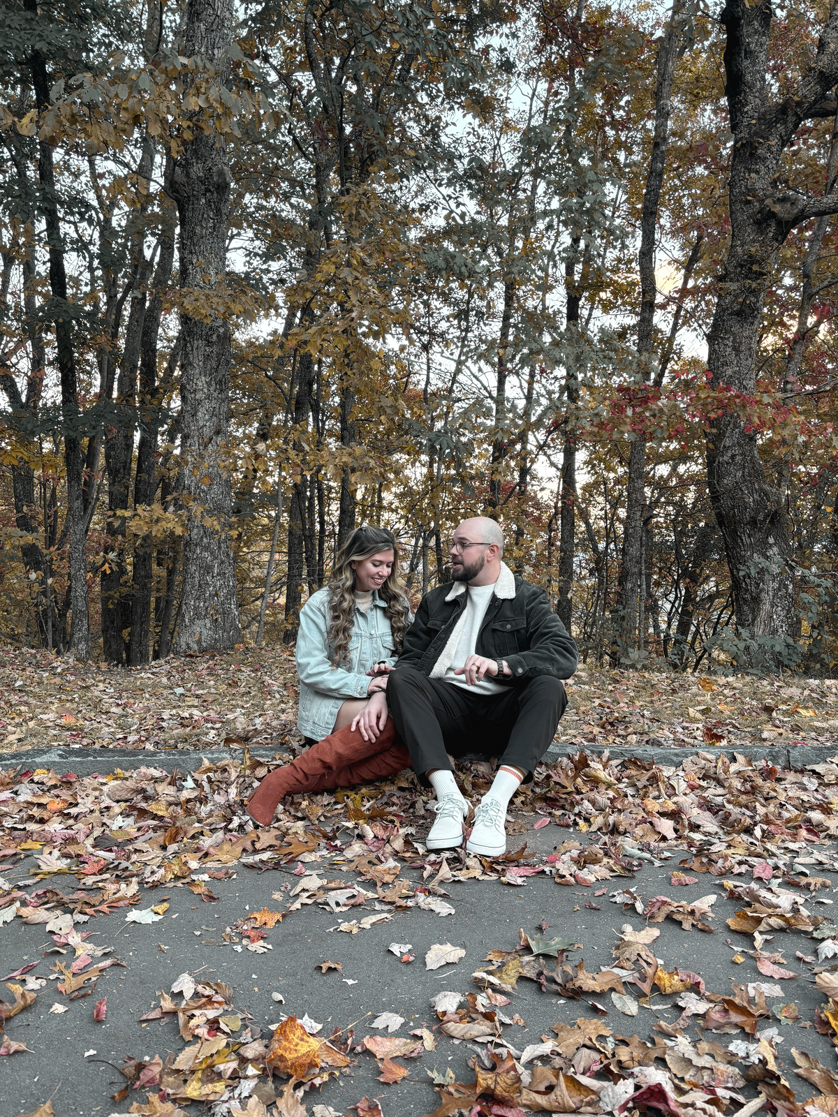 Sitting together on the edge of a path buried in fallen leaves, mid-conversation, bare autumn trees behind them.