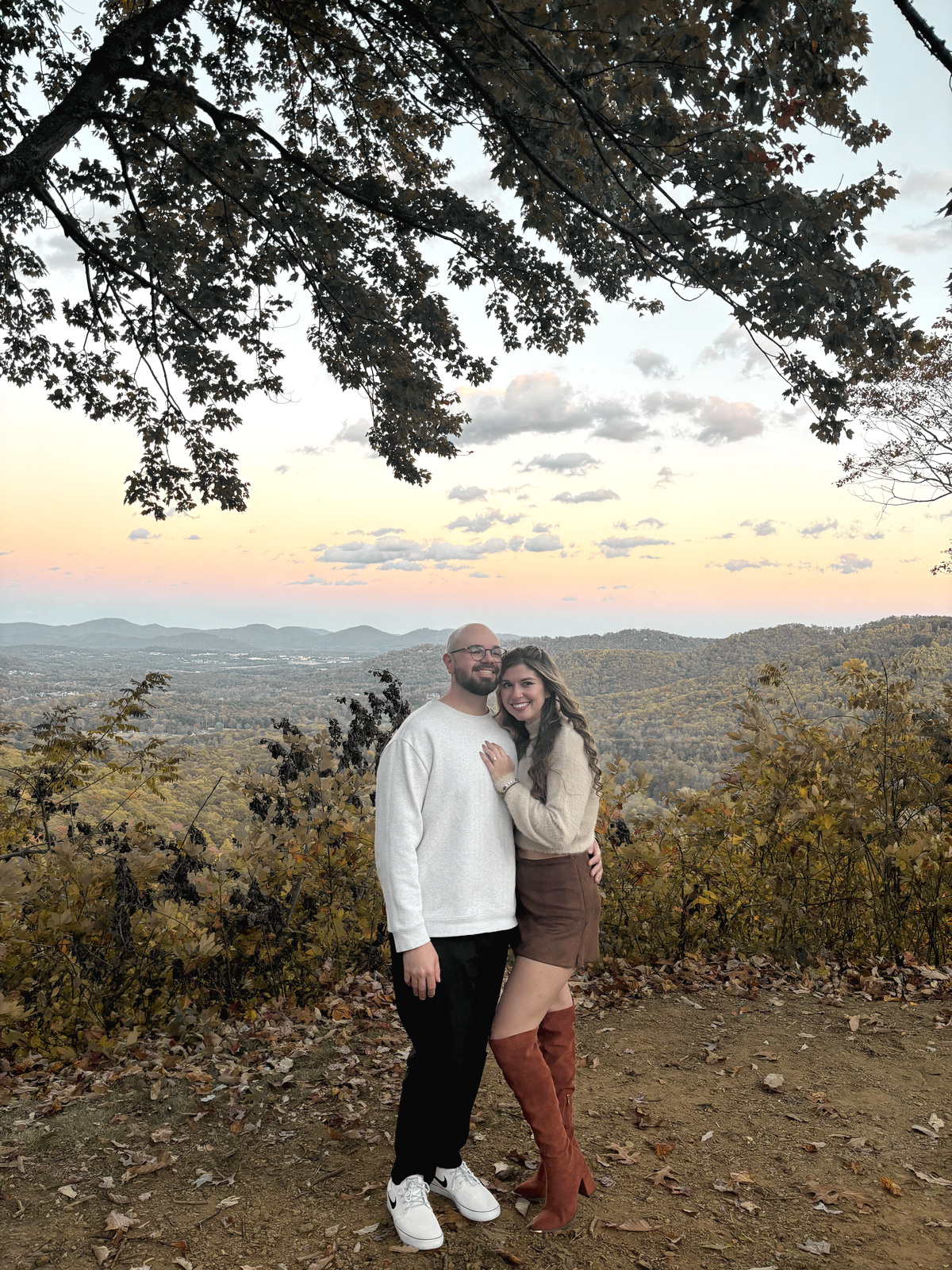 Nicole and Ian at a mountain overlook, arm in arm beneath a leafy branch, with a pale sunset over the ridges behind them.