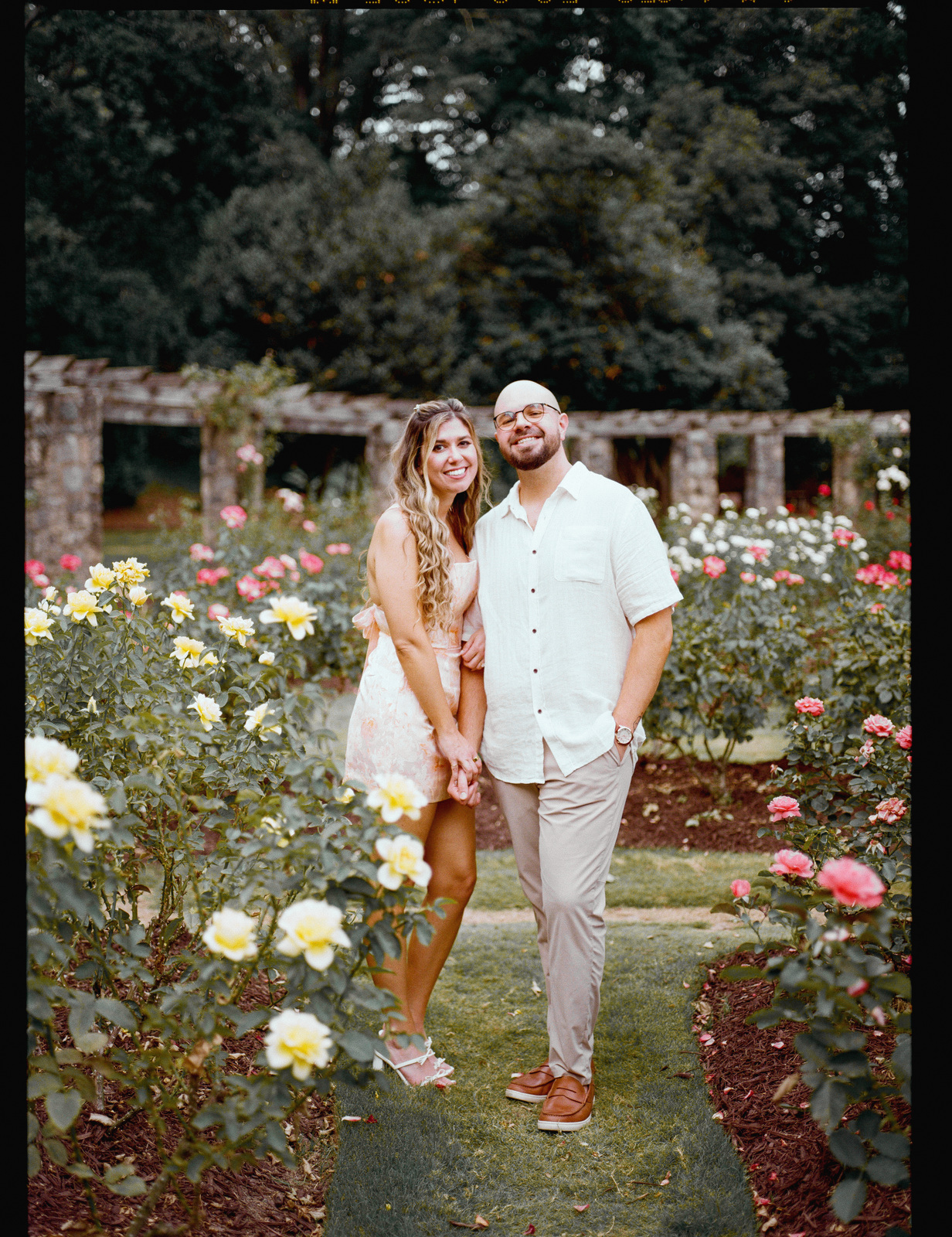 Standing together on the lawn between beds of yellow and pink roses, a long stone pergola behind them.
