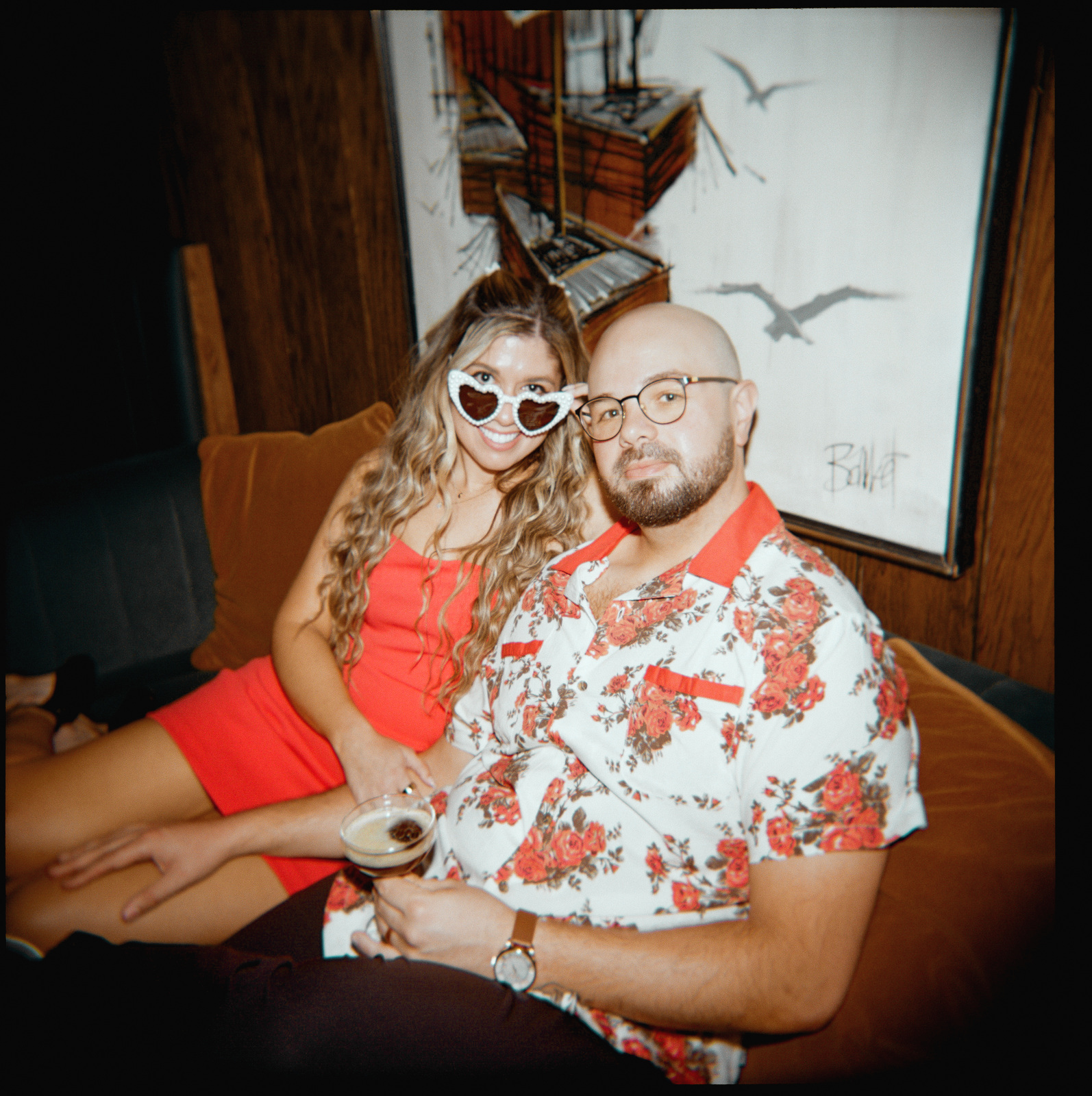 On a velvet couch beneath a painting, one wearing white heart-shaped sunglasses, the other holding a drink.