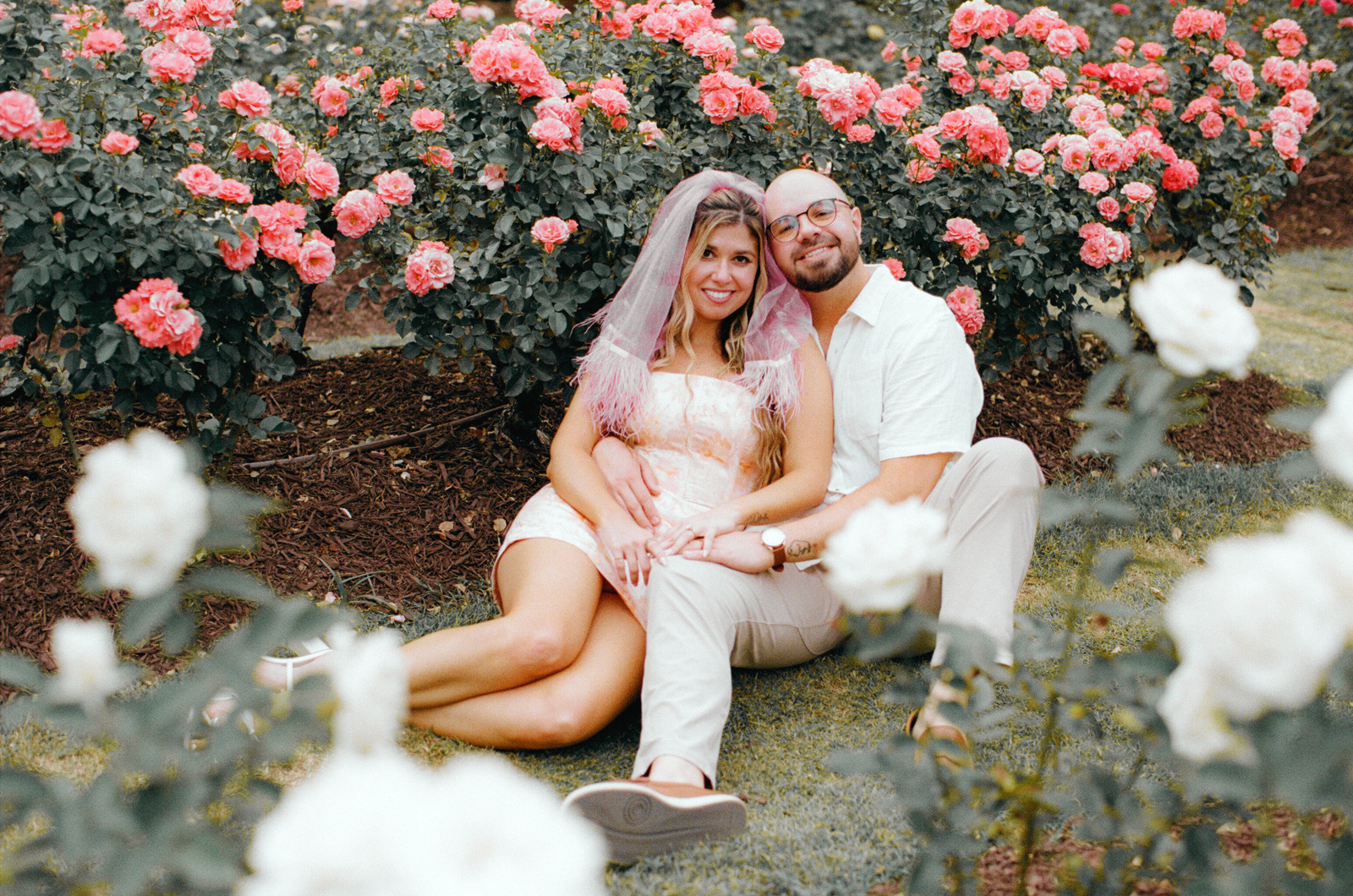 Sitting on the grass in front of a wall of coral roses, with white roses blurred in the foreground.