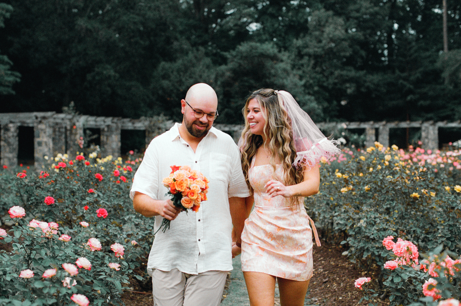 Walking hand in hand along a grass path through the rose beds, one carrying a bunch of orange roses, the other wearing a short pink veil.