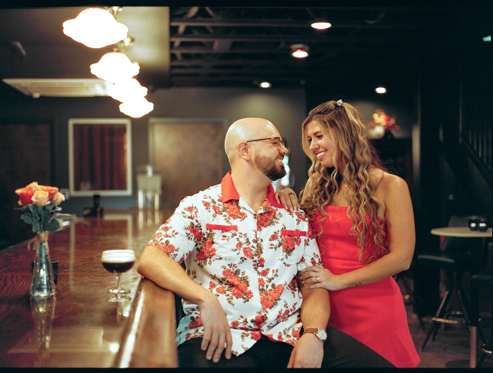 Leaning against a bar counter, laughing at each other, an espresso martini and a vase of roses beside them.