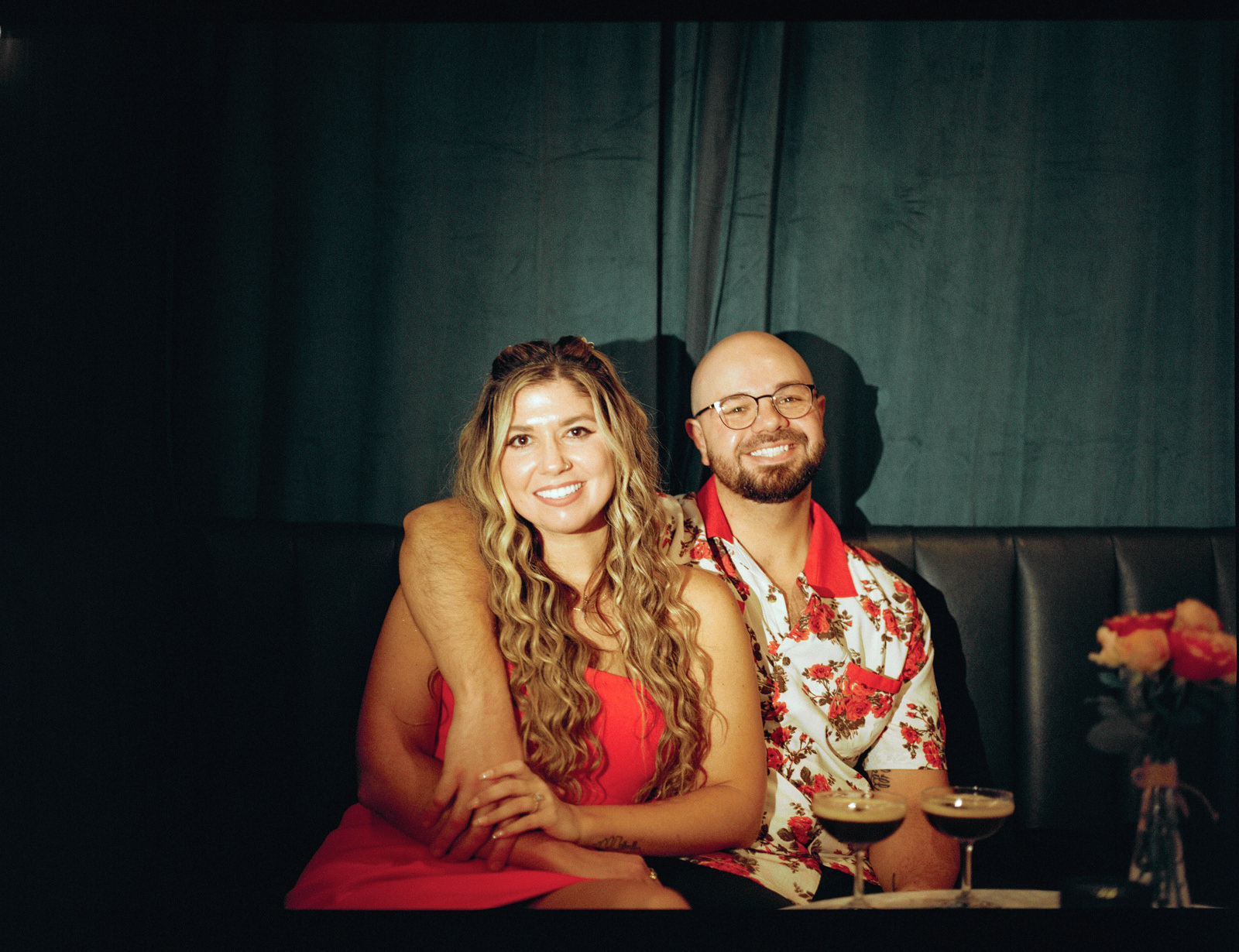 Side by side in a booth against a deep green curtain, two espresso martinis and a vase of roses on the table.