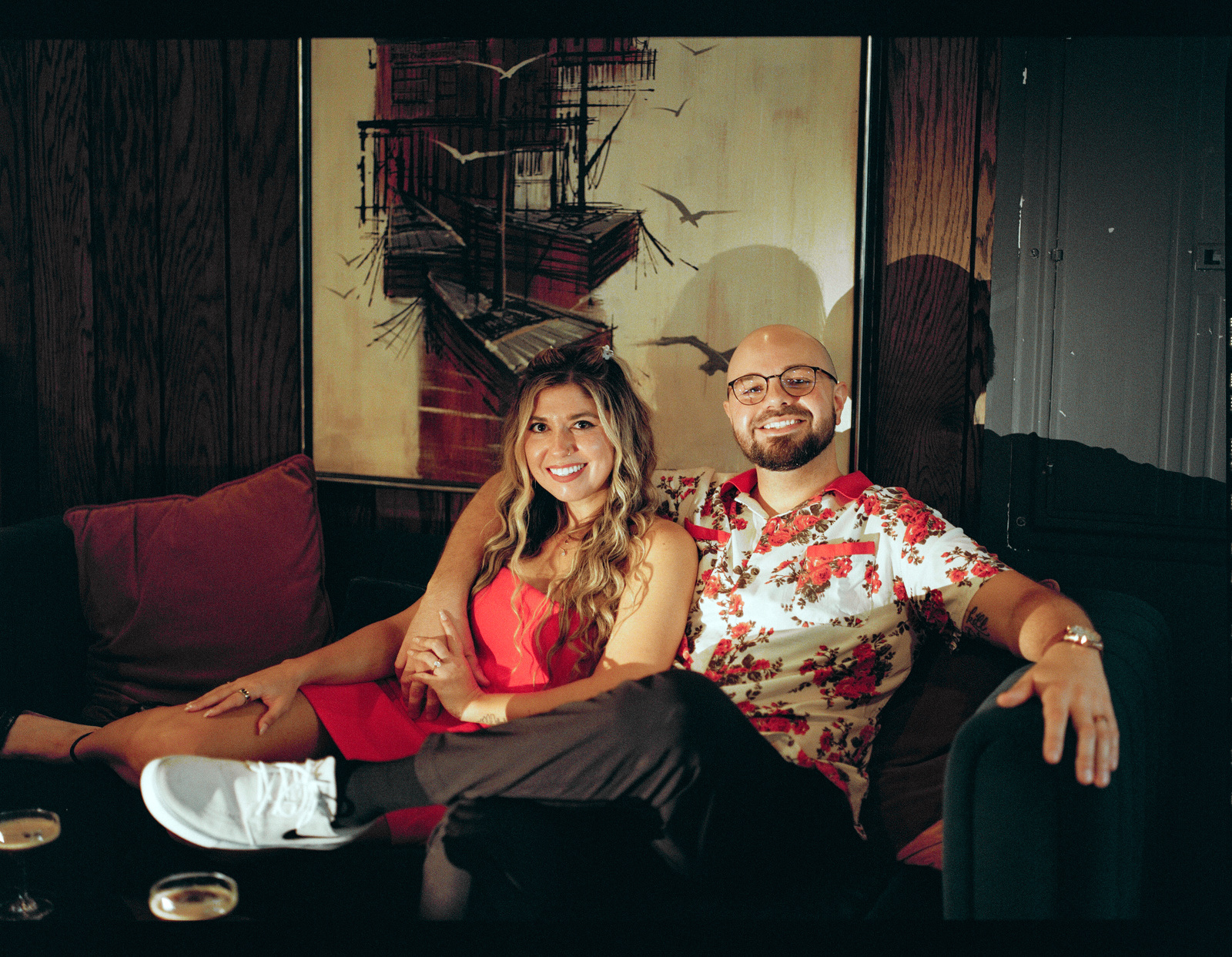 Settled into a red couch under a large painting, feet up, grinning at the camera.